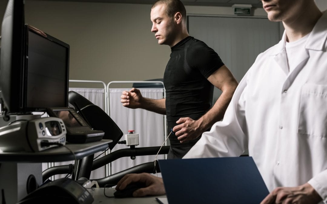 Photo of man in black shirt running on treadmill during cardiac stress test performed by doctor standing in the foreground